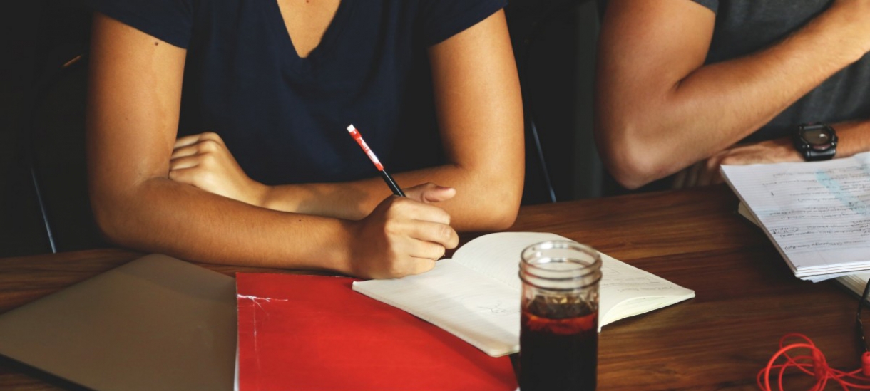 Individuals seated at a table with laptop, notepad, pencil, and drink