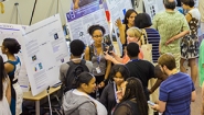 Students, faculty, and visitors gather around posters presenting student summer research.