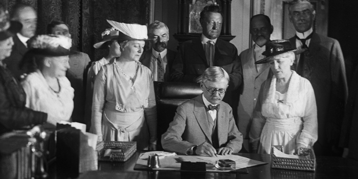 Cokie Roberts ’64 Reflects on Women’s Suffrage on the 100th Anniversary of the Senate Passing the 19th Amendment A black and white photo of a man signing a document with women standing behind him.