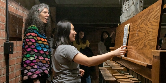 “All the Bells You Need”: Wellesley’s Carillon Reopens to Visitors after Renovation A student sits at the bench of a carillon with a teacher standing over her.