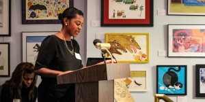 People of Color in Publishing Looks to Spark Change Across the Industry A woman speaks from a podium with artwork all around her