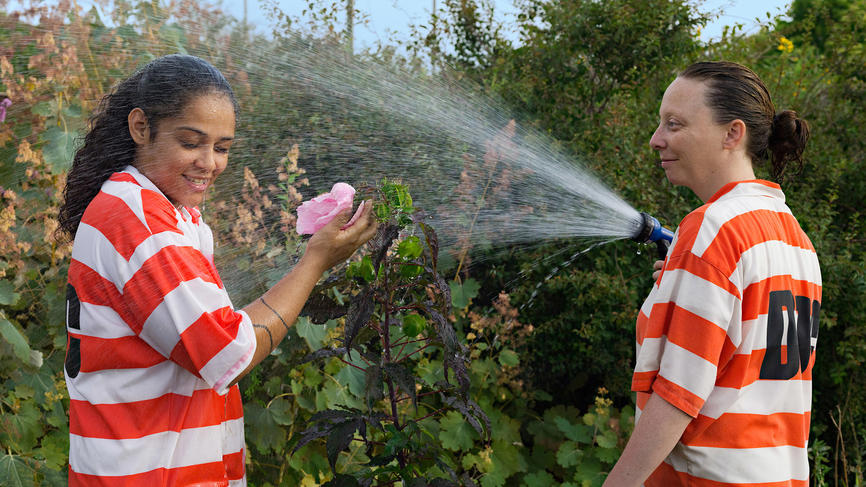 Two women in orange and white jumpsuits water flowers. The one holding the hose sprays the other woman.