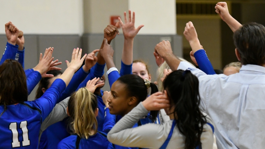 The Wellesley volleyball team stands with their hands up in a huddle.