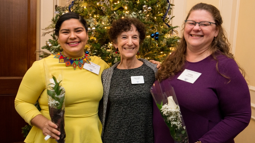 Three women stand in front of a Christmas tree which is decorated in yellow and purple.