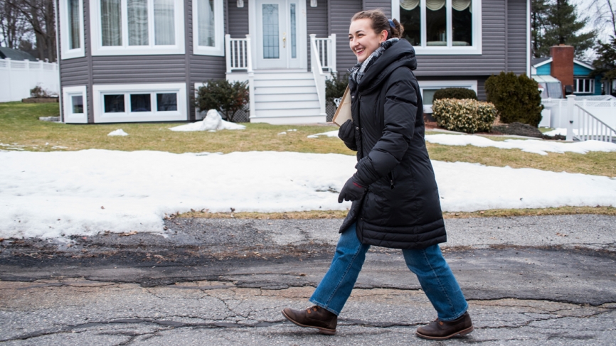 A woman walks down a snowy street in front of a gray house