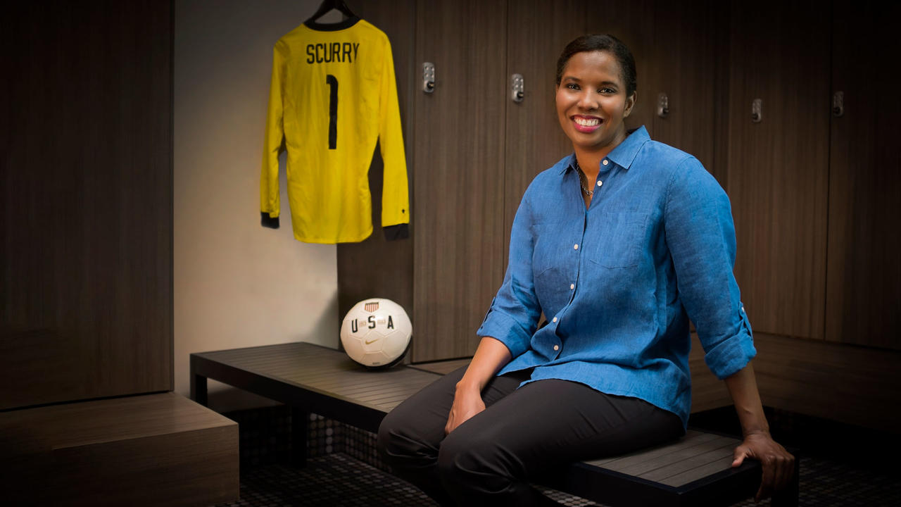 Briana Scurry posing on a bench with a soccer ball and jersey next to her