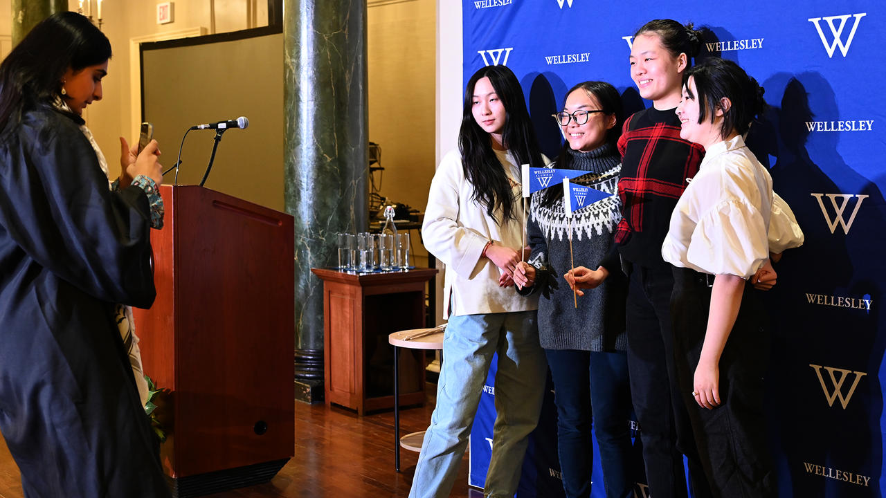 Four December graduates pose for a photo against a Wellesley logo backdrop.