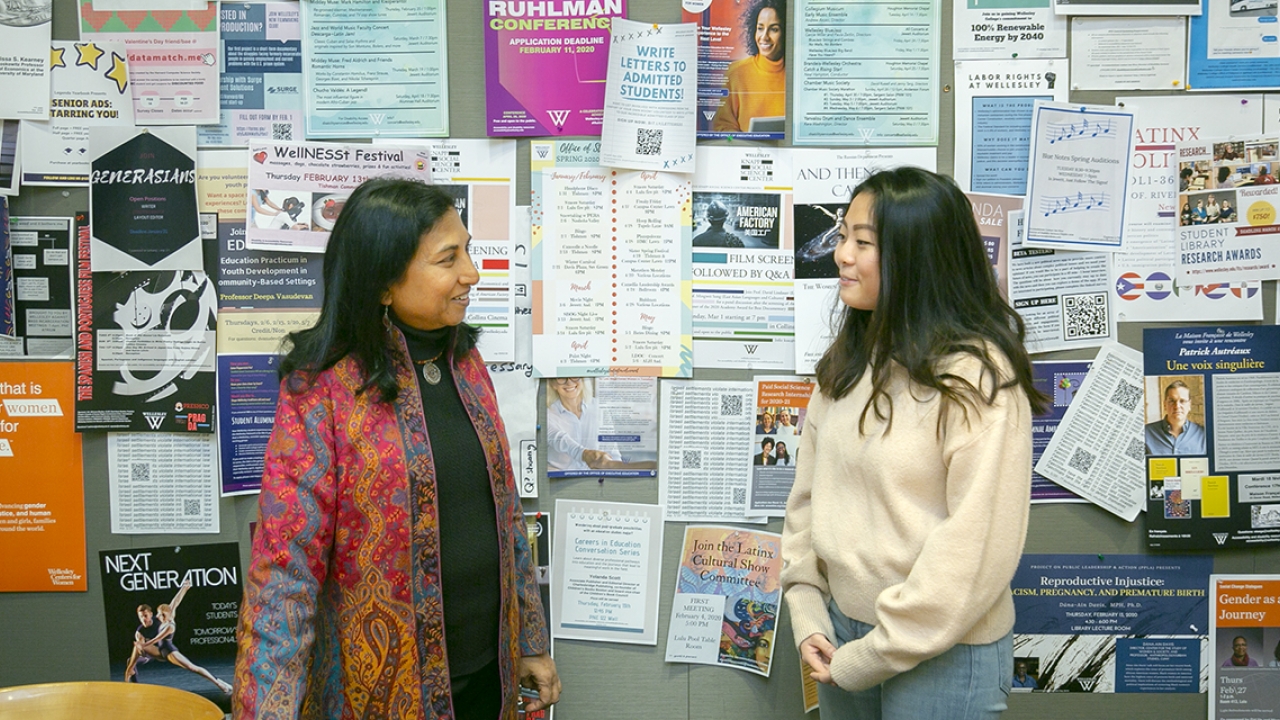 A student a guest speaker stand in front of a wall with posters