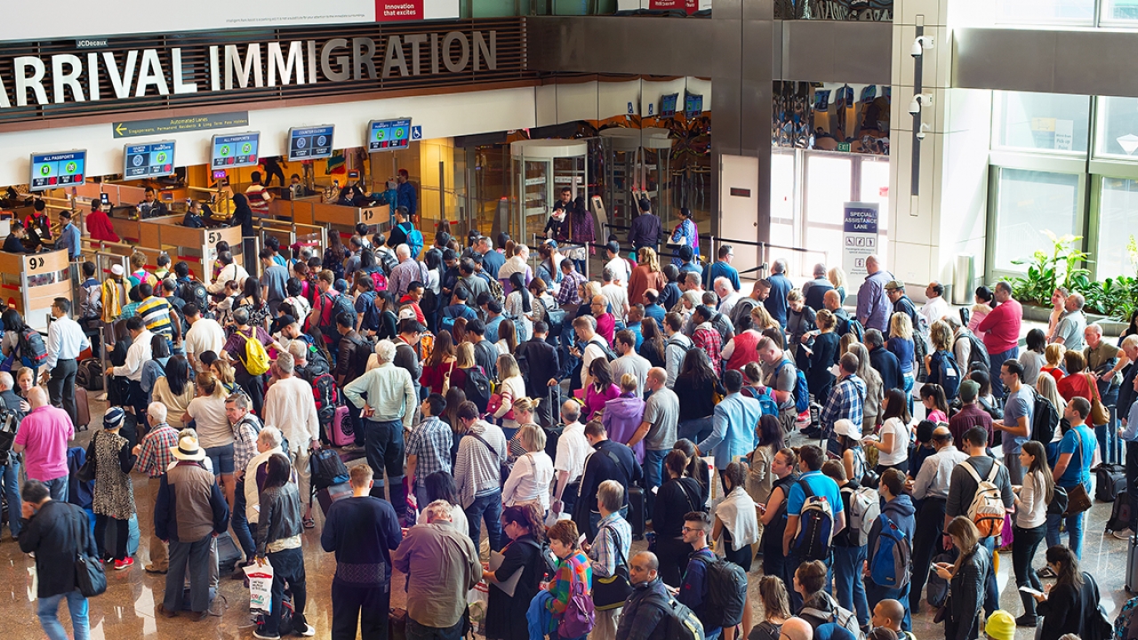 Hundreds of people stand in line at a transportation hub waiting to cross a boarder.