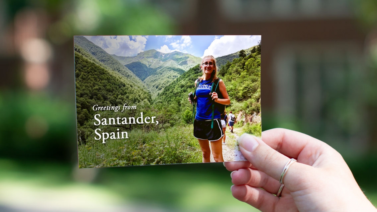 A student stands in a Wellesley shirt in front of a mountain range in Spain. Text reads, "Greetings from Santander, Spain"