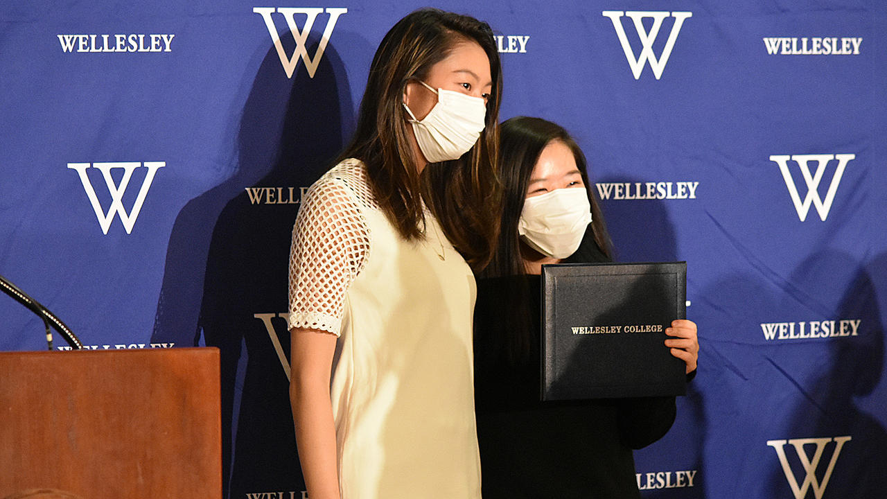 Two students pose for a photo with one student holding up her diploma case that read Wellesley College.