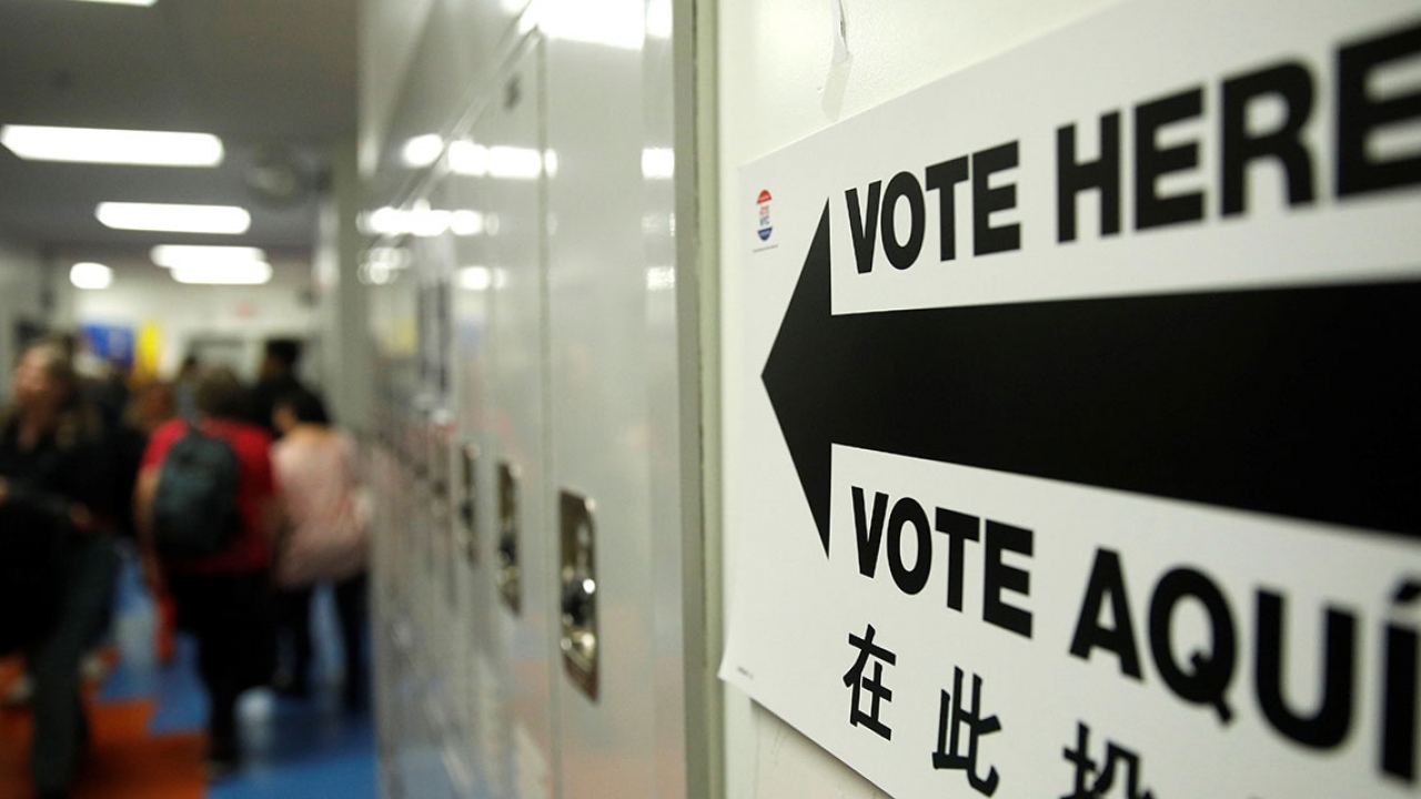 A sign on a wall by lockers reads Vote Here in English and Spanish