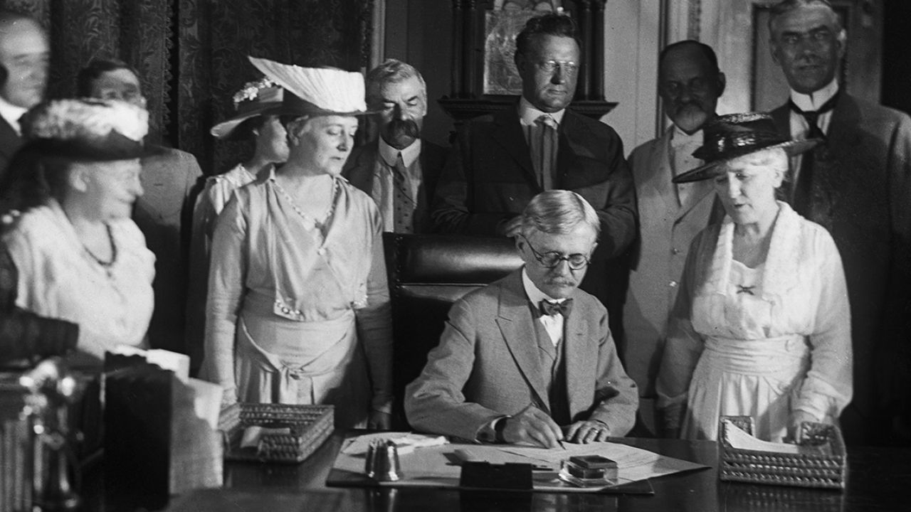 A black and white photo of a man signing a document with women standing behind him.