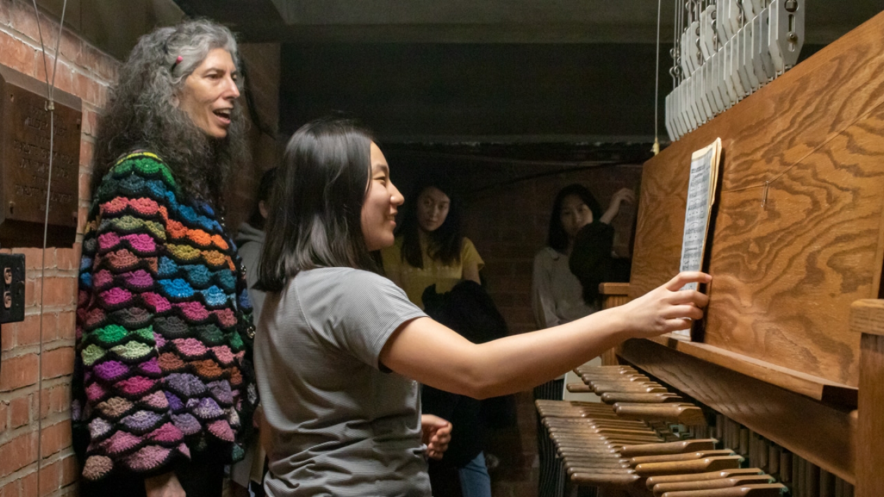 A student sits at the bench of a carillon with a teacher standing over her.