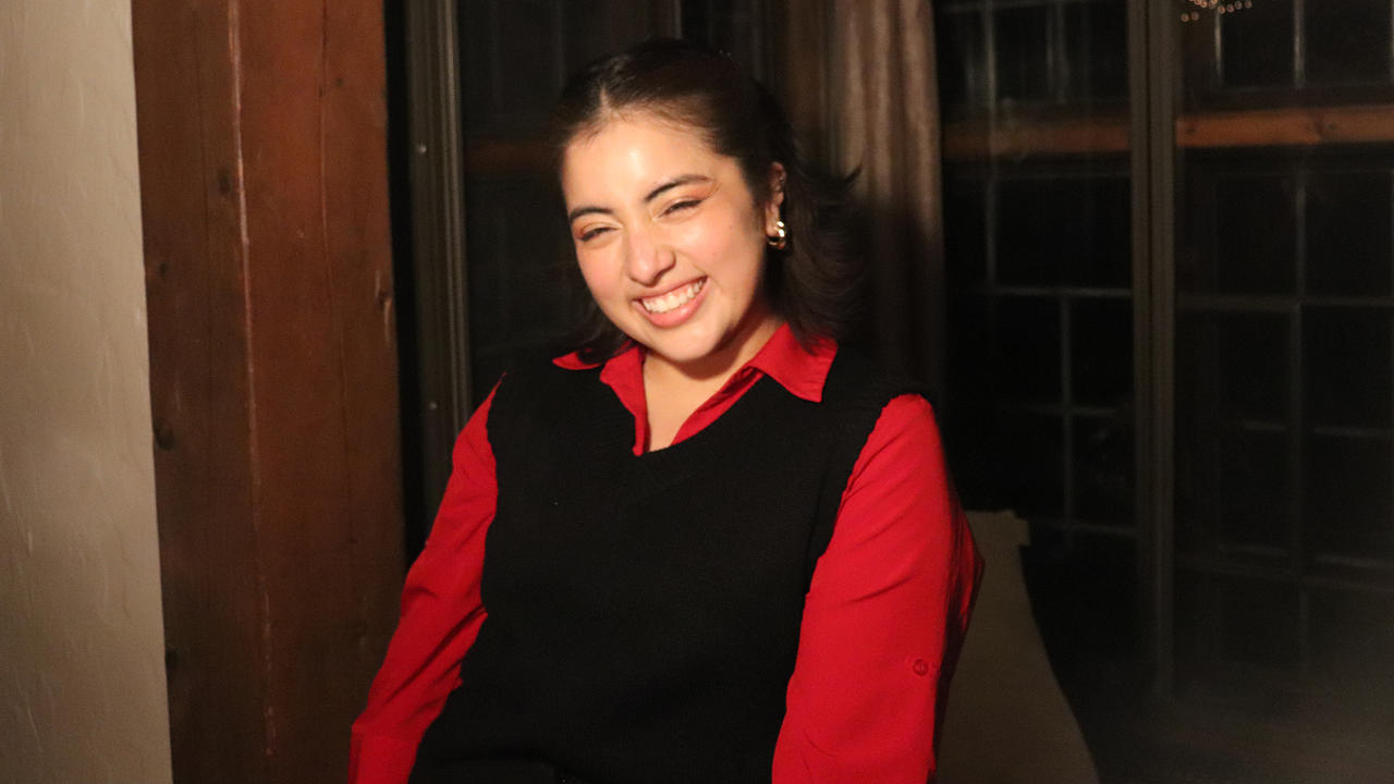 Lizette Ortega wears a long-sleeved red shirt and black sweater vest. She smiles at the camera.