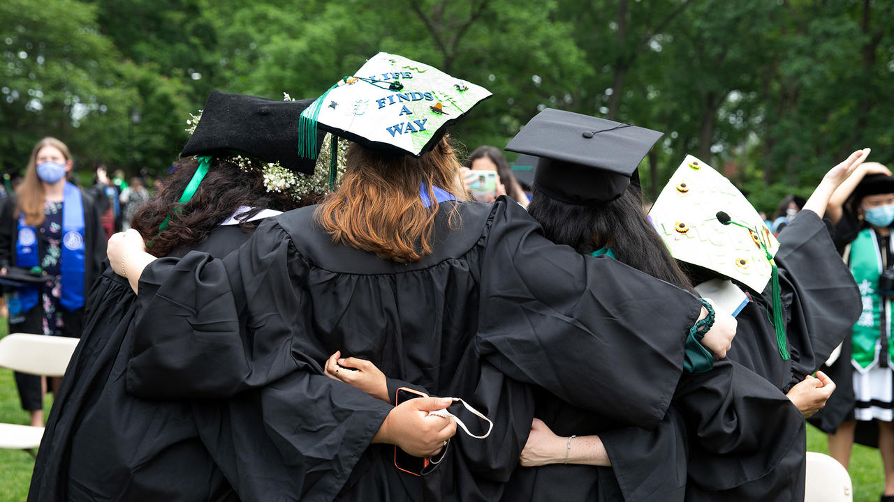 The backs of four class of 2021 graduates that have their arms around each other. A mortarboards reads Life Finds A Way