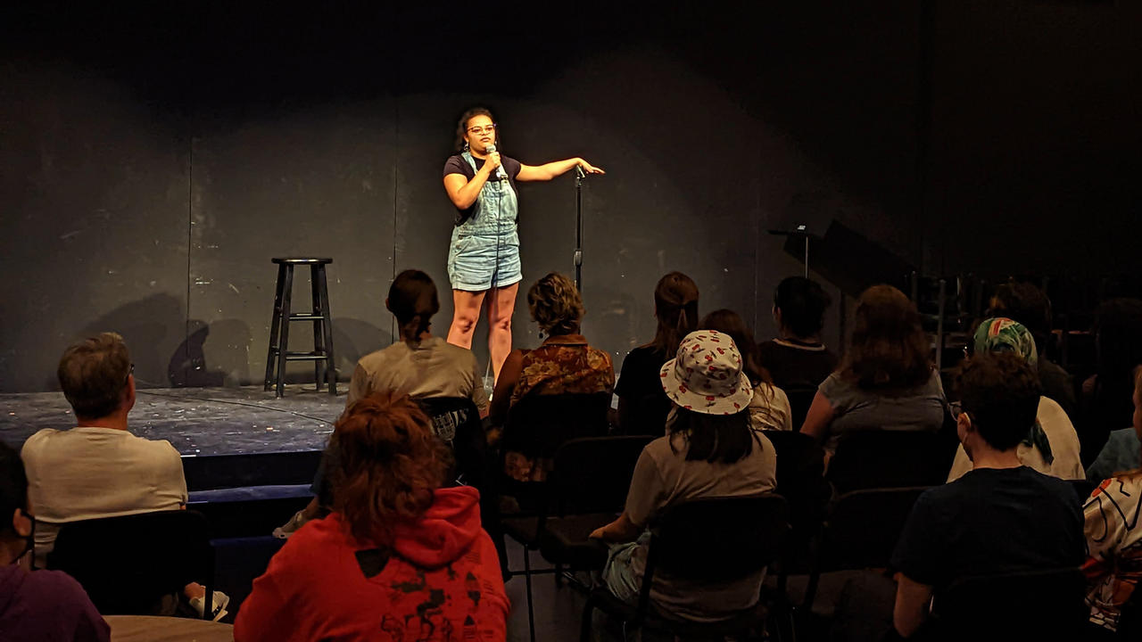 A student stands in the spotlight on stage, performing stand-up comedy for an audience.